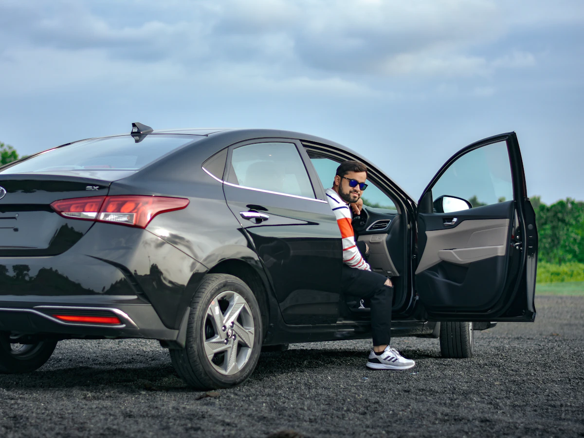 A driver sitting in the open door of a black sedan on a sunny day